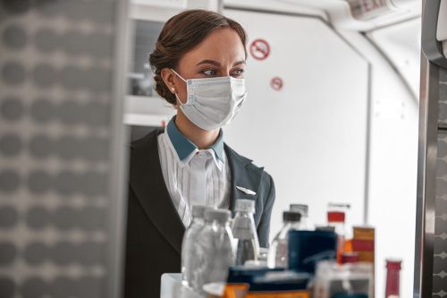 Stewardess carrying food trolley in corridor of airplane jet. Interior of modern plane. European woman wear uniform, latex gloves and medical mask. Civil aviation. Air travel concept