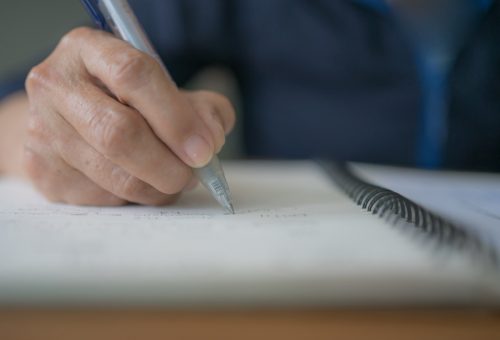 Senior woman wrinkle hand holds pen handwriting diary, letter or the will on a notebook. Female pensioner drafts idea of testament. Aged journalist writing report, memos or documenting information.