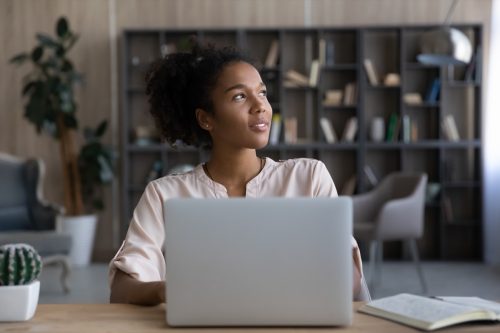 Dreamy African American woman looking to aside, distracted from laptop, sitting at work desk.