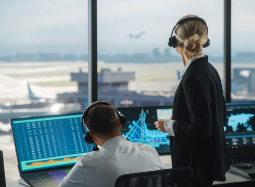 Female and Male Air Traffic Controllers with Headsets Talk in Airport Tower. Office Room is Full of Desktop Computer Displays with Navigation Screens, Airplane Departure and Arrival Data for the Team.