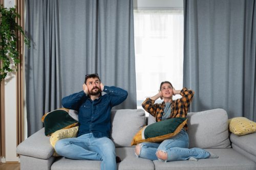 Young couple is sitting on a sofa in their apartment looking up and holding their hands to plug their ears