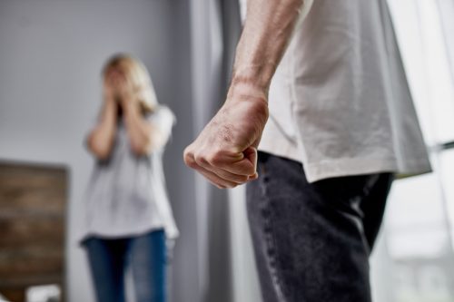 photo of male fist, angry man and scared woman in the background, she close her face with hands. 