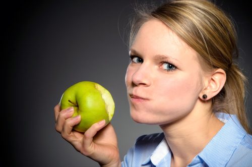 Young blond woman eats joyfully a green apple.