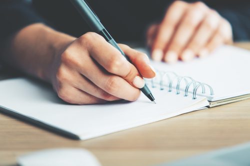 Close up of woman's hands writing in spiral notepad placed on wooden desktop with various items