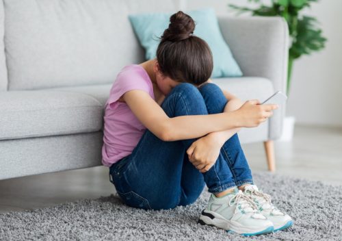 Depressed teen girl with cellphone crying on floor at home.