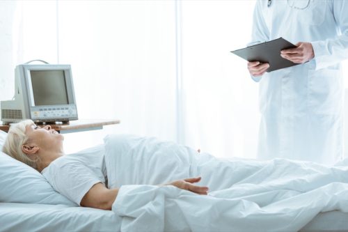 Doctor with clipboard and sick senior woman lying on bed.