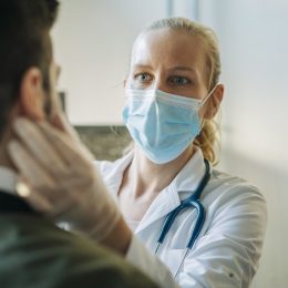 Doctor wearing surgical mask examining infected patient. Mature healthcare worker checking mid adult man. They are at pharmacy.