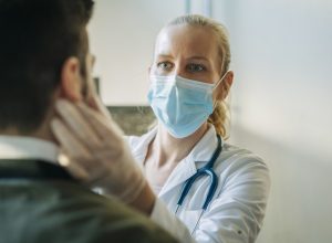 Doctor wearing surgical mask examining infected patient. Mature healthcare worker checking mid adult man. They are at pharmacy.