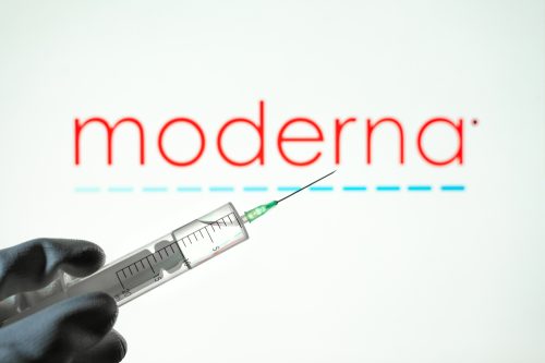 A gloved hand holding a syringe of vaccine or a booster in front of the Moderna logo