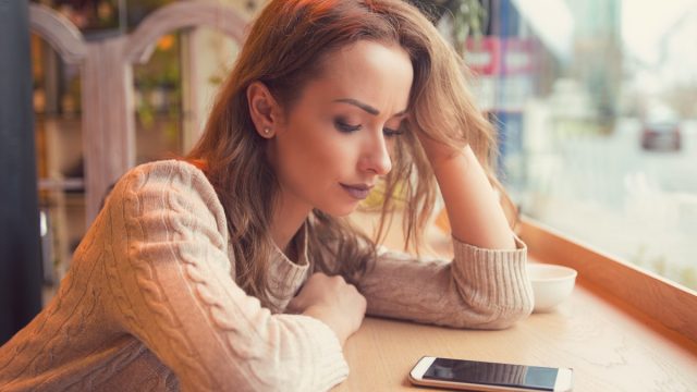 Young woman sitting at table in cafe looking at phone being unhappy with breakup.