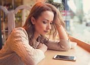 Young woman sitting at table in cafe looking at phone being unhappy with breakup.