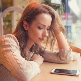Young woman sitting at table in cafe looking at phone being unhappy with breakup.