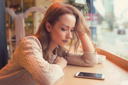 Young woman sitting at table in cafe looking at phone being unhappy with breakup.