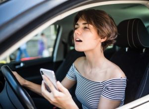 Closeup portrait, young woman driving in black car and checking her phone, annoyed by navigation gps system or bad text message or email, isolated outdoors background