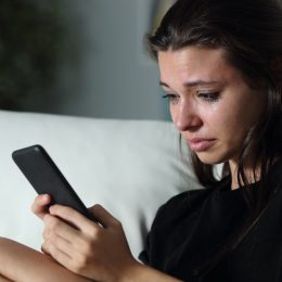 Sad teenage girl checking smart phone cyber bullying messages sitting on a couch in the dark at home.