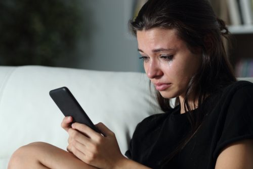 Sad teenage girl checking smart phone cyber bullying messages sitting on a couch in the dark at home.