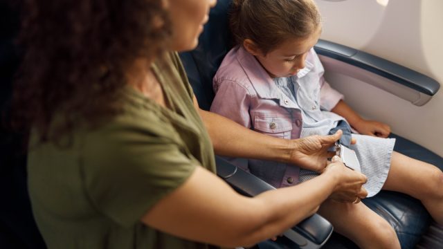 Mother helping her little daughter to adjust and tight seatbelt on an airplane for safe flight. Traveling by airplane during Covid19, family concept