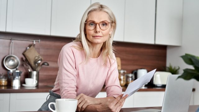 Middle aged woman wearing glasses holding document, working on laptop sitting at home office kitchen