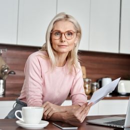 Middle aged woman wearing glasses holding document, working on laptop sitting at home office kitchen