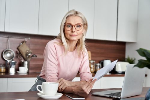 Middle aged woman wearing glasses holding document, working on laptop sitting at home office kitchen