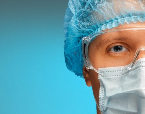 Young man doctor in cap, glasses and mask looks at camera, close-up
