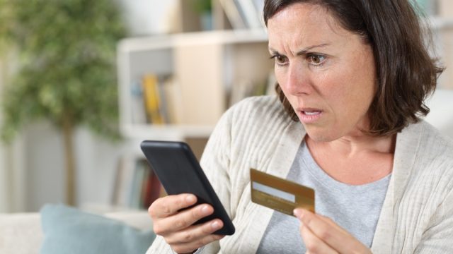 Worried adult woman paying online on smart phone with credit card sitting on the sofa at home