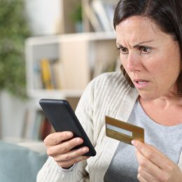 Worried adult woman paying online on smart phone with credit card sitting on the sofa at home