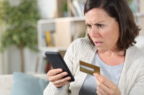 Worried adult woman paying online on smart phone with credit card sitting on the sofa at home