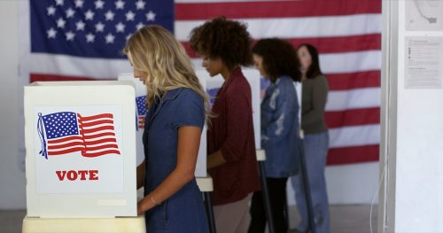 Four women of various demographics, young blonde woman in front, filling in ballots and casting votes in booths at polling station, US flag on wall at back. Focus on booth signage