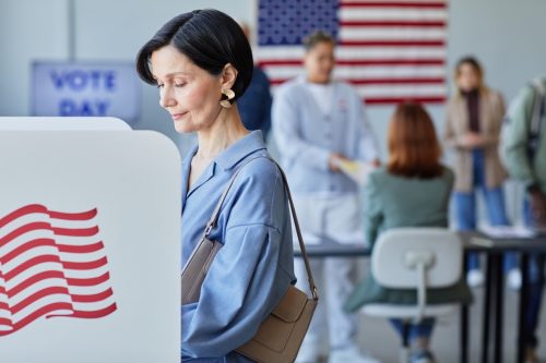 Side view portrait of elegant adult woman voting in booth on election day with American flags in shot, copy space