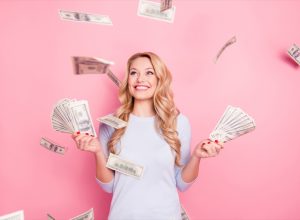 Portrait of pretty charming positive cute successful lucky cheerful girl standing under shower from money having a lot of money in hands isolated on pink background