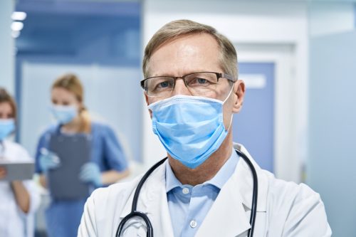 Mature old medical healthcare professional doctor wearing white coat, stethoscope, glasses and face mask standing in hospital looking at camera.