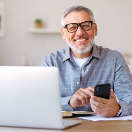 Handsome smiling senior man wearing glasses using mobile phone while sitting at his cozy workplace with laptop at home, retired male chatting with friends in social media, typing on smartphone