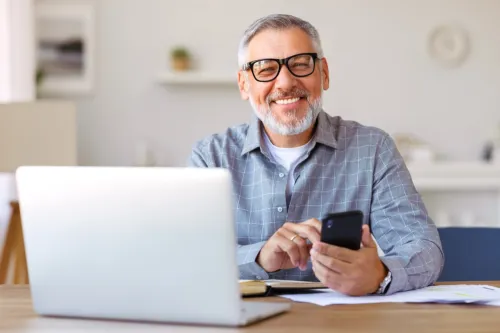 Handsome smiling senior man wearing glasses using mobile phone while sitting at his cozy workplace with laptop at home, retired male chatting with friends in social media, typing on smartphone