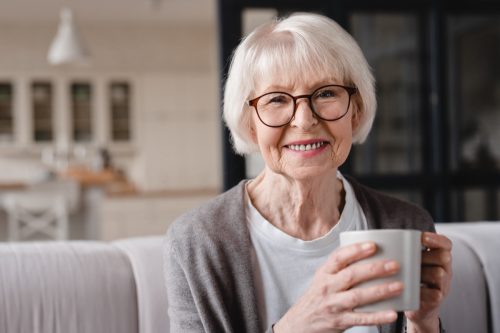 Comfort relaxed senior old elderly woman grandmother drinking hot beverage tea coffee at home looking at camera in the living room.