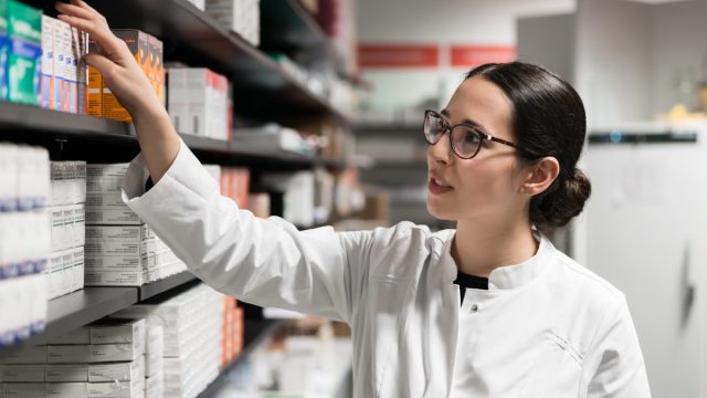 Portrait of a dedicated female pharmacist taking a medicine from the shelf, while wearing eyeglasses and lab coat during work in a modern drugstore with various pharmaceutical products.