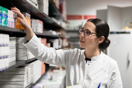 Portrait of a dedicated female pharmacist taking a medicine from the shelf, while wearing eyeglasses and lab coat during work in a modern drugstore with various pharmaceutical products.