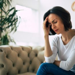 Close-up photo of a young woman, who is sitting on a sofa with her eyes closed, touching her head while suffering from a migraine.