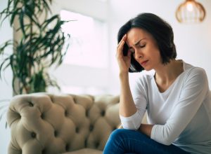 Close-up photo of a young woman, who is sitting on a sofa with her eyes closed, touching her head while suffering from a migraine.
