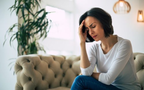 Close-up photo of a young woman, who is sitting on a sofa with her eyes closed, touching her head while suffering from a migraine.