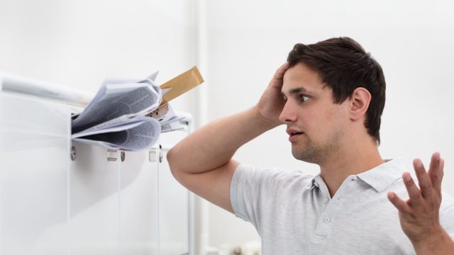 Close-up of man with hand on head in front of overloaded mailbox with junk mail