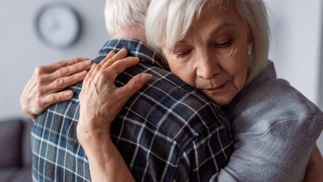 Senior woman with closed eyes embracing husband sick on dementia.