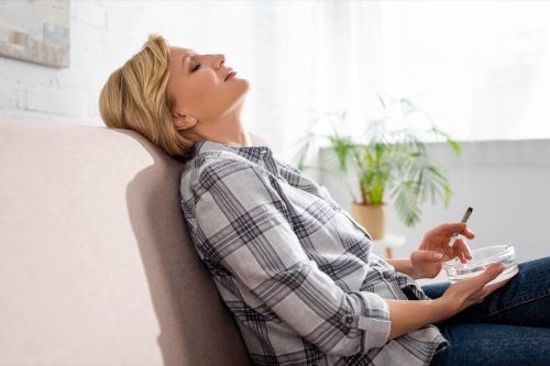 Mature woman with closed eyes sitting on sofa and holding joint with marijuana.