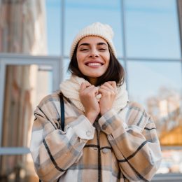 Smiling woman walking in street in winter outfit with coffee wearing checkered coat, white knitted hat and scarf.