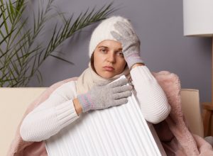 Portrait of sick unhealthy woman in coat and hat sit in cold living room with radiator, has no heating in her house, suffering flu and headache, looking at camera with sad expression.