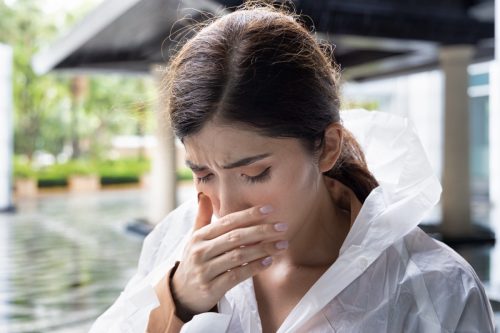 Unwell woman wearing raincoat feeling sick, catching cold with runny nose, nasal congestion and fever during rain fall in rainy monsoon season