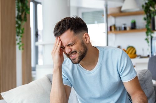 Closeup of young man suffering from headache at home, touching his temples, copy space, blurred background. Migraine, headache, stress, tension problem, hangover concept