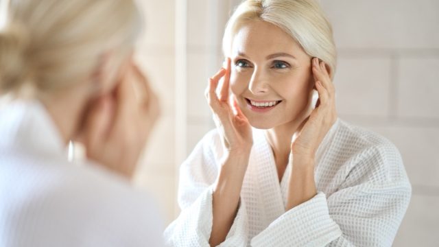 Mature woman standing in bathroom after shower touching face, looking at reflection in mirror smiling doing morning beauty routine.