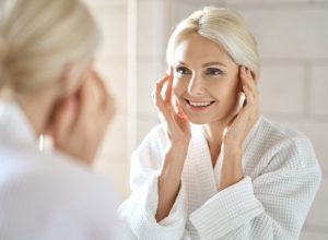 Mature woman standing in bathroom after shower touching face, looking at reflection in mirror smiling doing morning beauty routine.