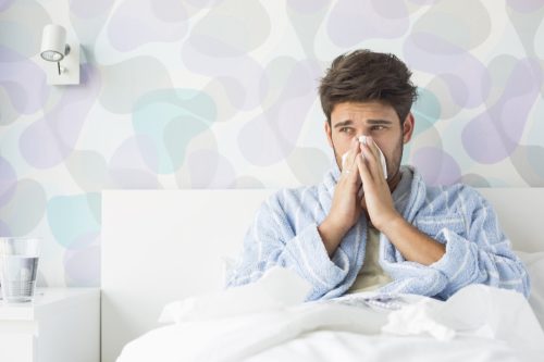 Sick man blowing his nose while sitting on bed at home